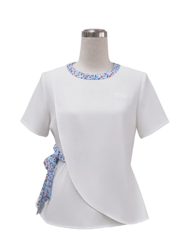 Women's white short-sleeve corporate tunic top featuring an asymmetric crossover hem, blue floral collar trim, and a matching side-tie ribbon designed by Modoleen Singapore for healthcare, corporate, and receptionist uniform programs.